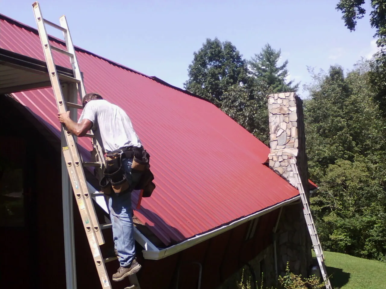Roofer on a ladder beside a red metal roof and stone chimney — Soffit & Fascia Repair by Roofing Brothers in Moravian Falls
